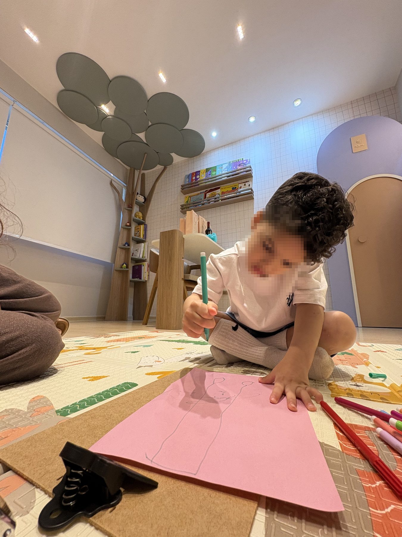 Criança desenhando no tapete da sala infantil, com a estante em árvore ao fundo